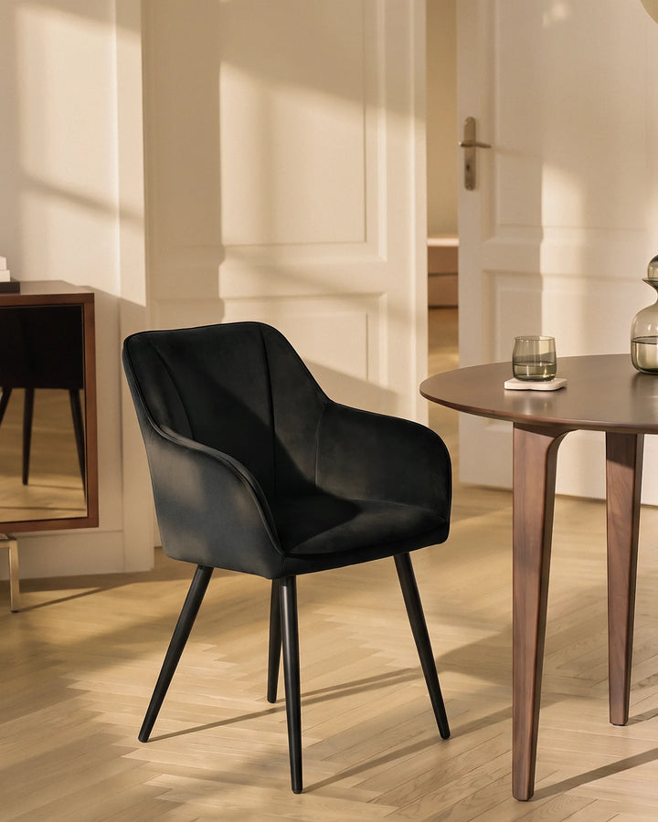 Earven black velvet dining chair styled beside a wooden dining table in a warm neutral dining space with framed wall art and full-length curtains