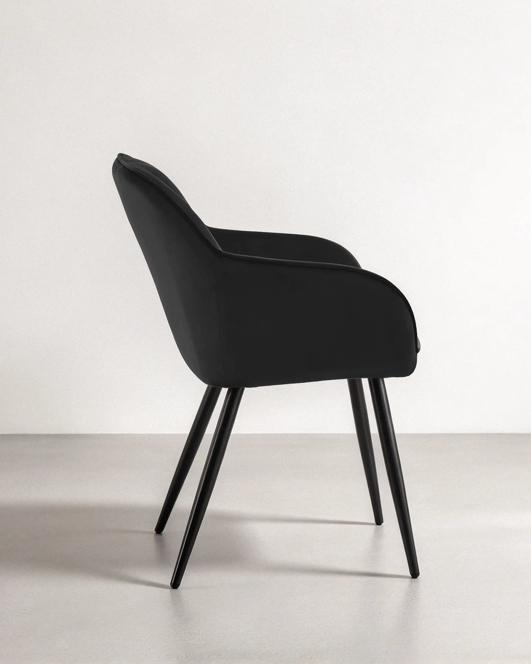 Side profile of the Earven black velvet dining chair highlighting its wraparound backrest, supportive armrests, cushioned seat and slim black legs