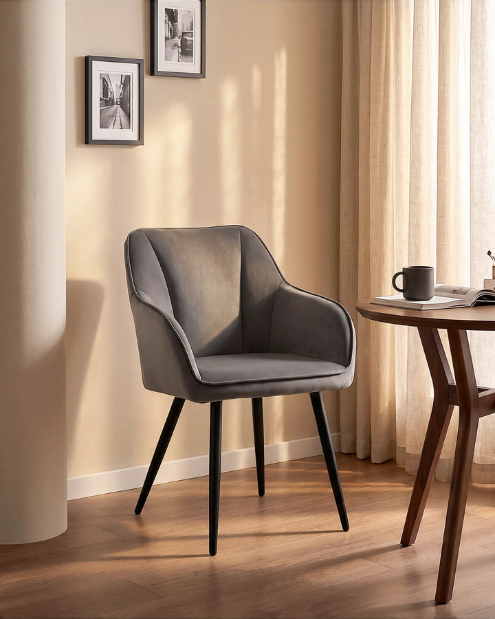 Earven grey velvet dining chair styled beside a wooden dining table in a warm neutral dining space with framed wall art and full-length curtains