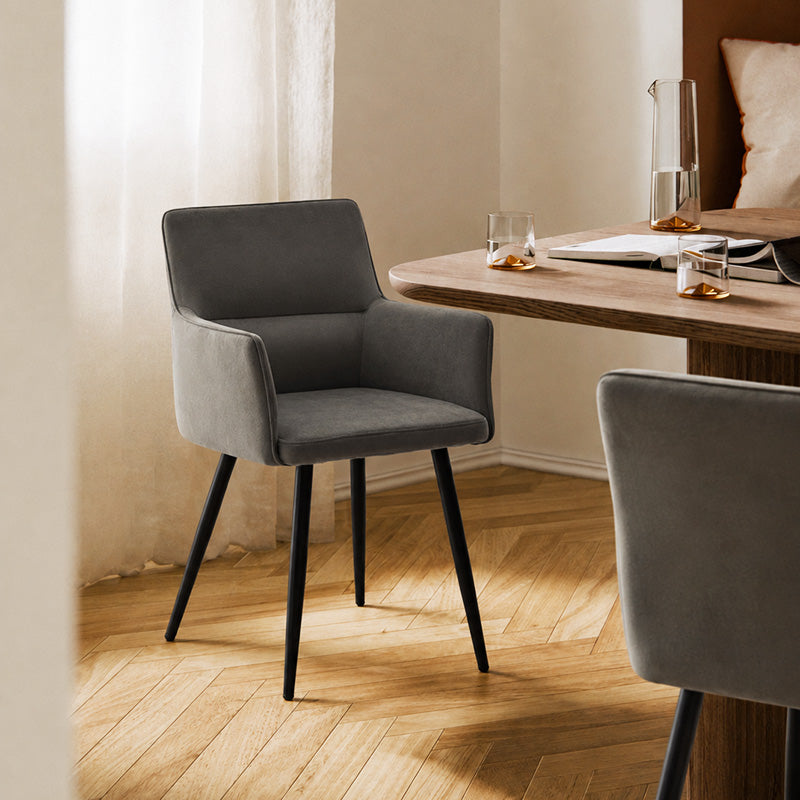 Ebern grey velvet dining chair with upholstered armrests and black metal legs, styled beside a wooden dining table in a modern dining room.