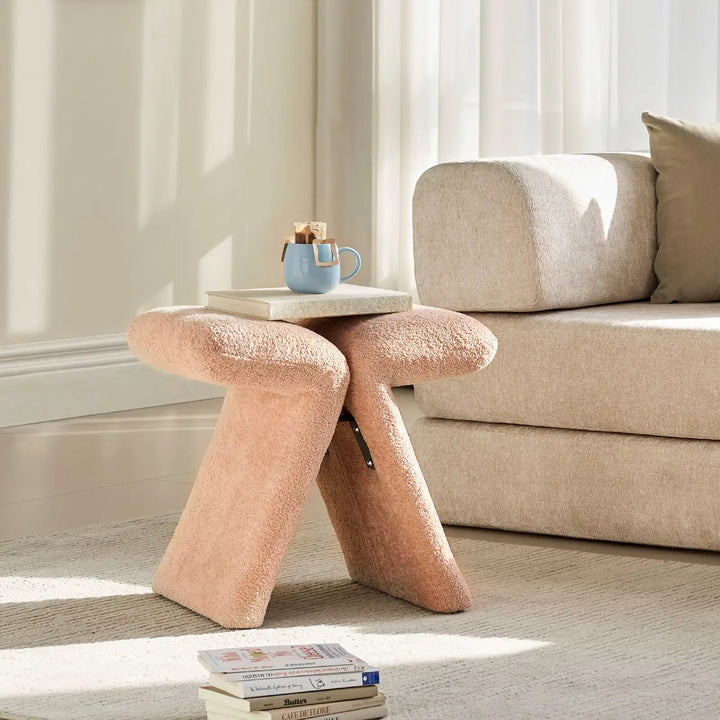 Faye Warm Blush Accent Stool styled beside a sofa in a modern living room, used as a side stool with books and cup of coffee