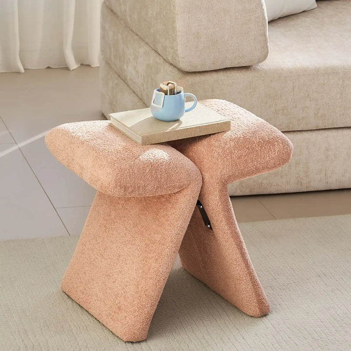 Faye Warm Blush Accent Stool placed by the window next to a sofa in a contemporary living room
