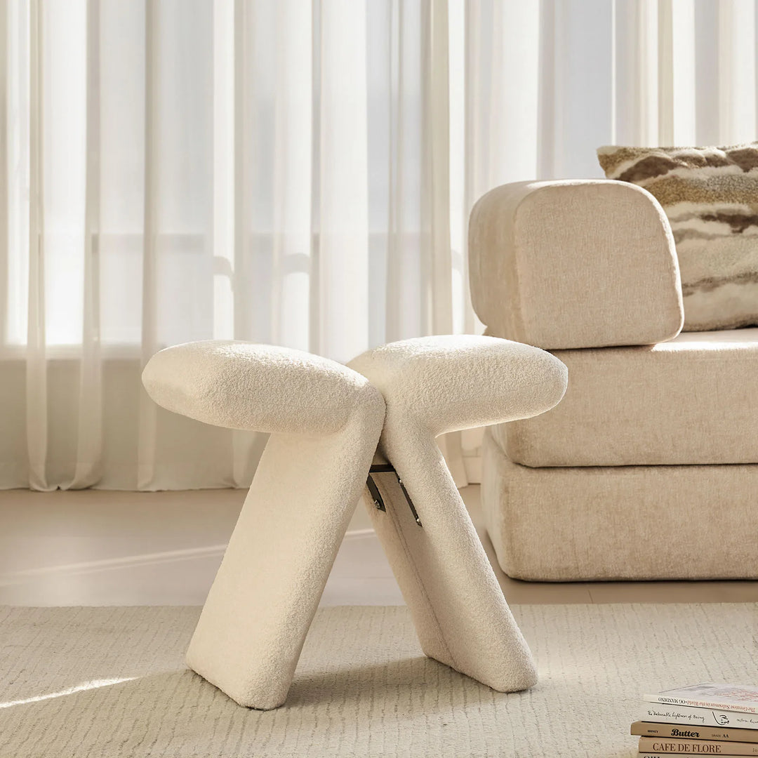 Faye warm white Accent Stool placed by the window next to a sofa in a contemporary living room.