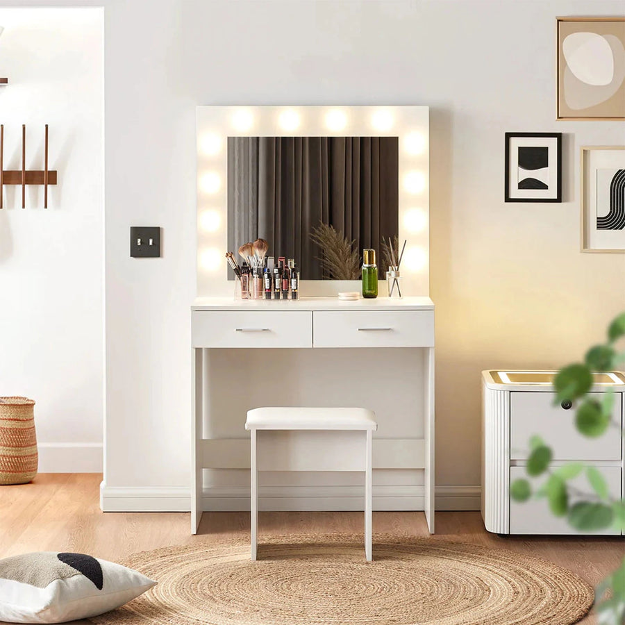 A Hollywood-style white vanity table with a mirror surrounded by bright bulb lights and a cushioned stool, placed in a modern bedroom with a bedside table beside it.