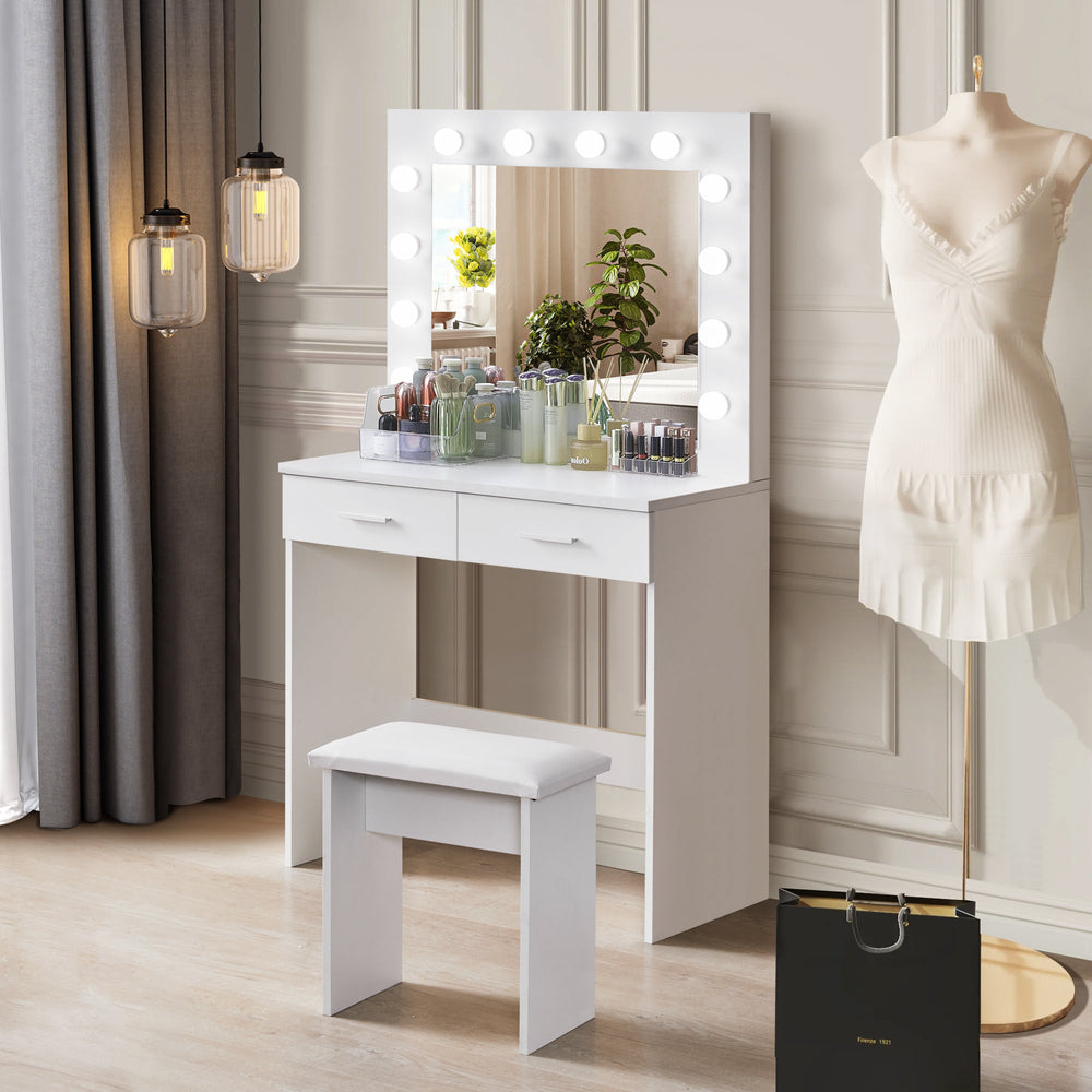 A Hollywood-style white vanity table with a mirror surrounded by bright bulb lights and a cushioned stool, placed in a modern bedroom with a clothes hanger beside it.