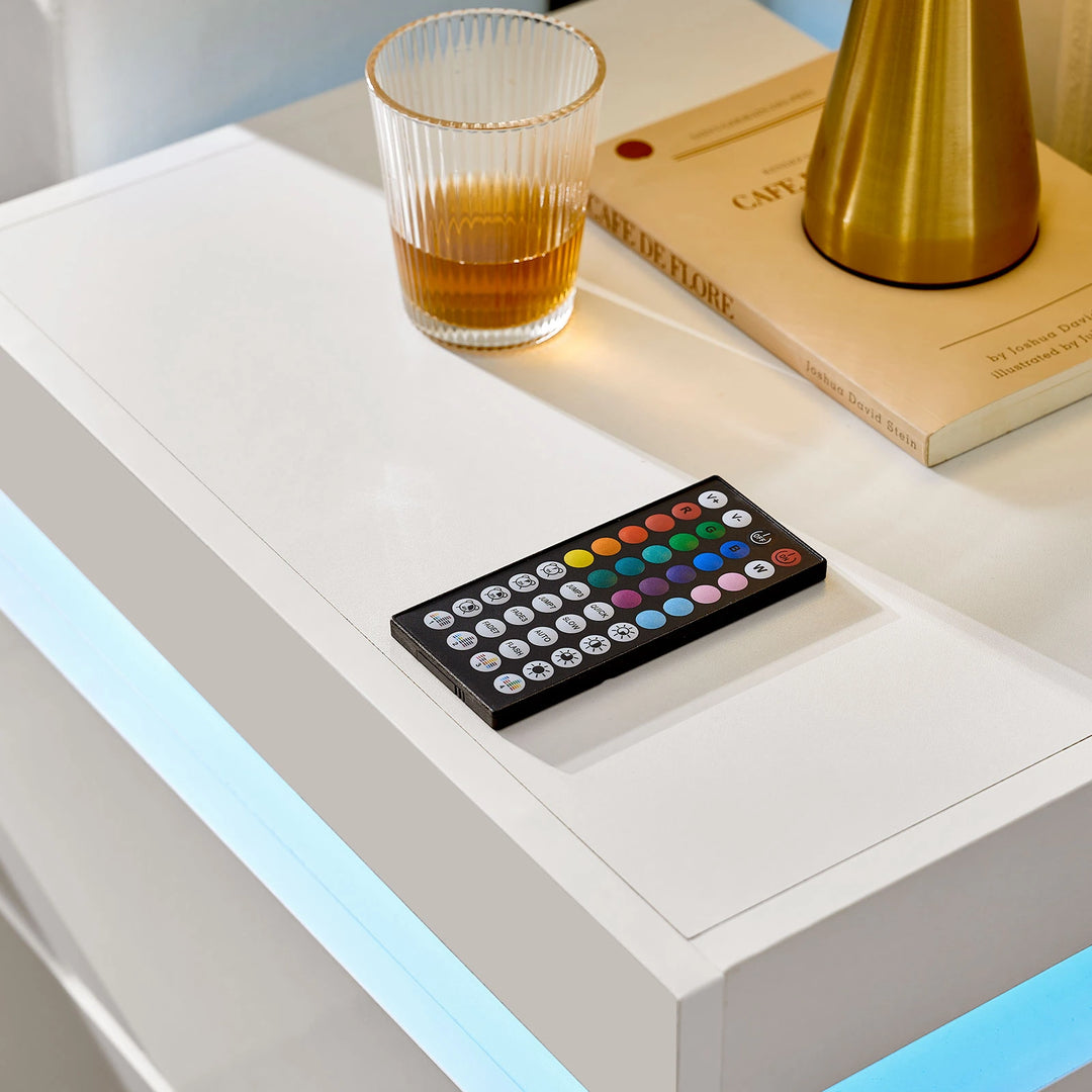 The close view of the Kumar Bedside Table's table top, with a remote controller, a glass, a book and a lamp on it, in a bright room.