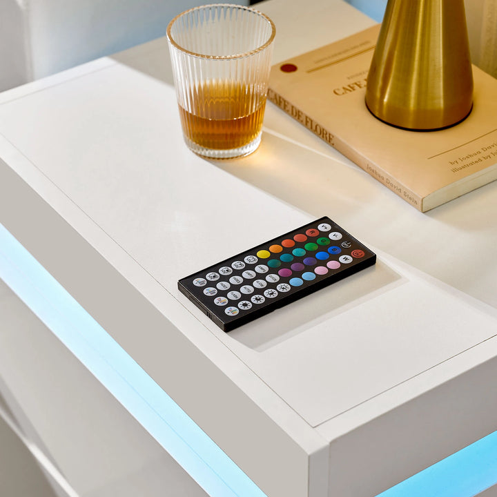 The close view of the Kumar Bedside Table's table top, with a remote controller, a glass, a book and a lamp on it, in a bright room.