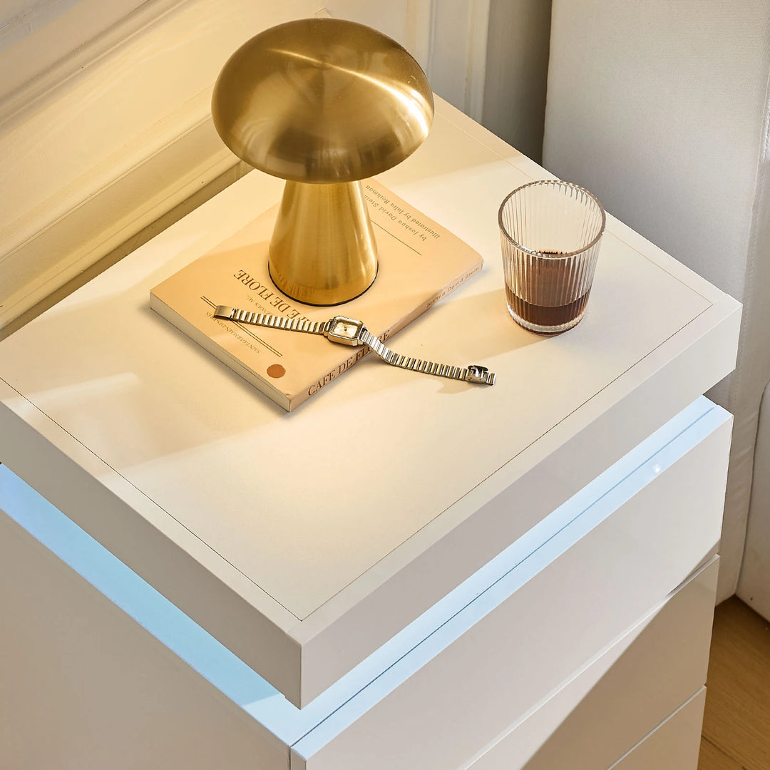 The close view of the Kumar Bedside Table's table top, with a glass, a watch, a book and a lamp on it, in a bright room.