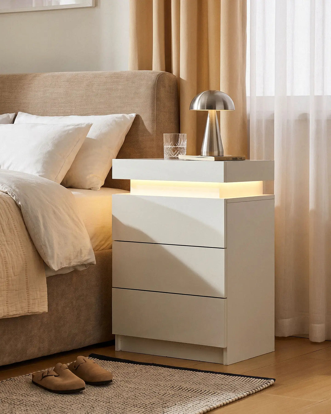 White high gloss smart bedside table with 3 drawers and warm LED light in a bedroom.