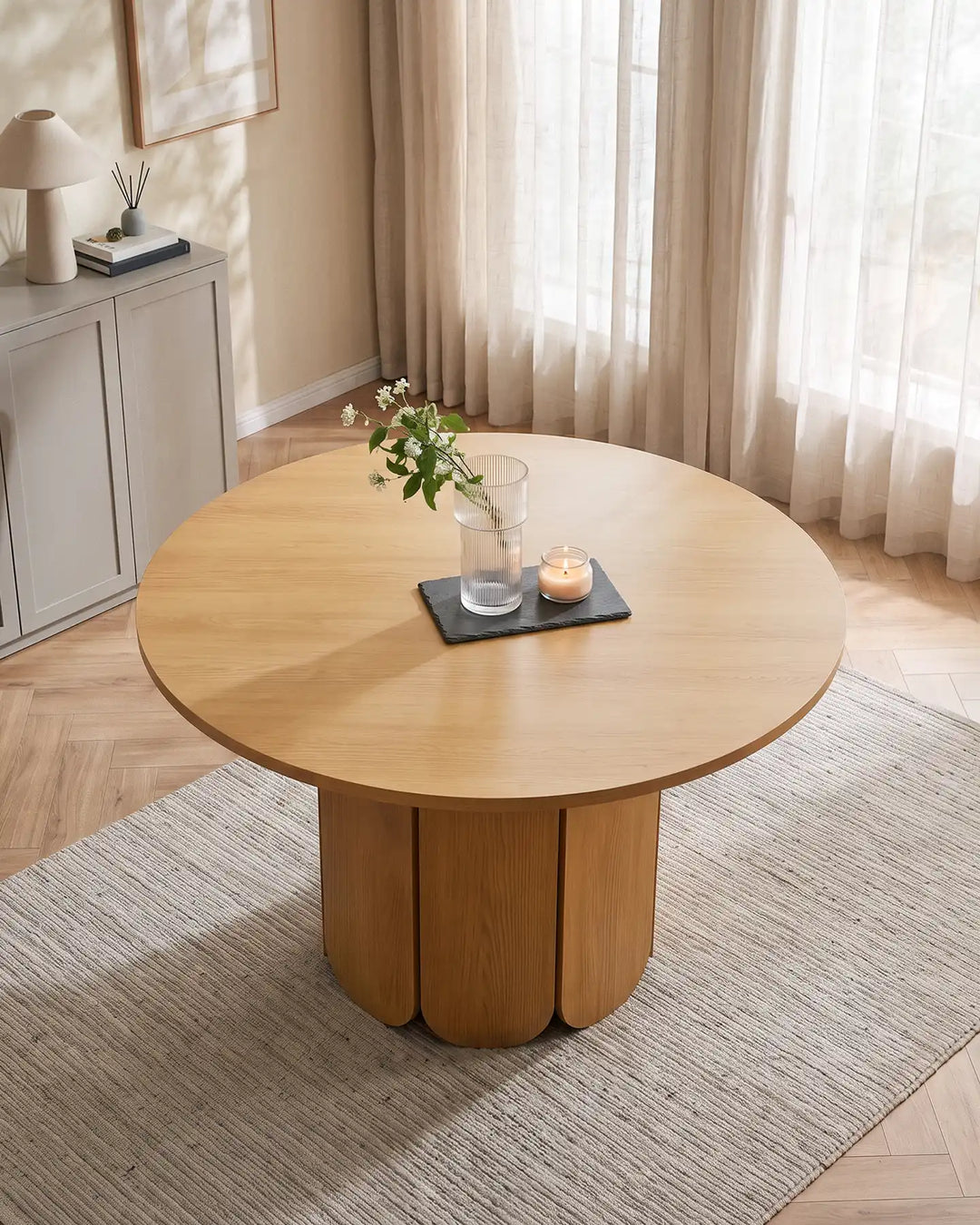 Nara round dining table in a natural oak-effect finish, styled with a small vase and candle tray in a warm neutral dining room with sheer curtains and a textured rug