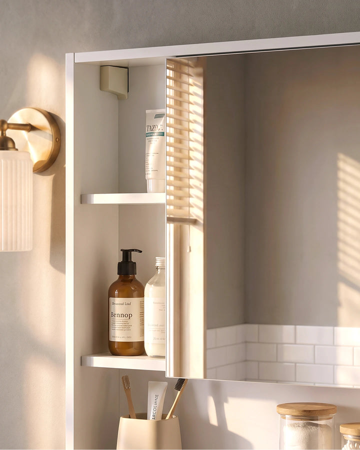 Close-up of a white bathroom mirror cabinet with a mirrored door, open side shelves and warm brass lighting in a modern bathroom setting.
