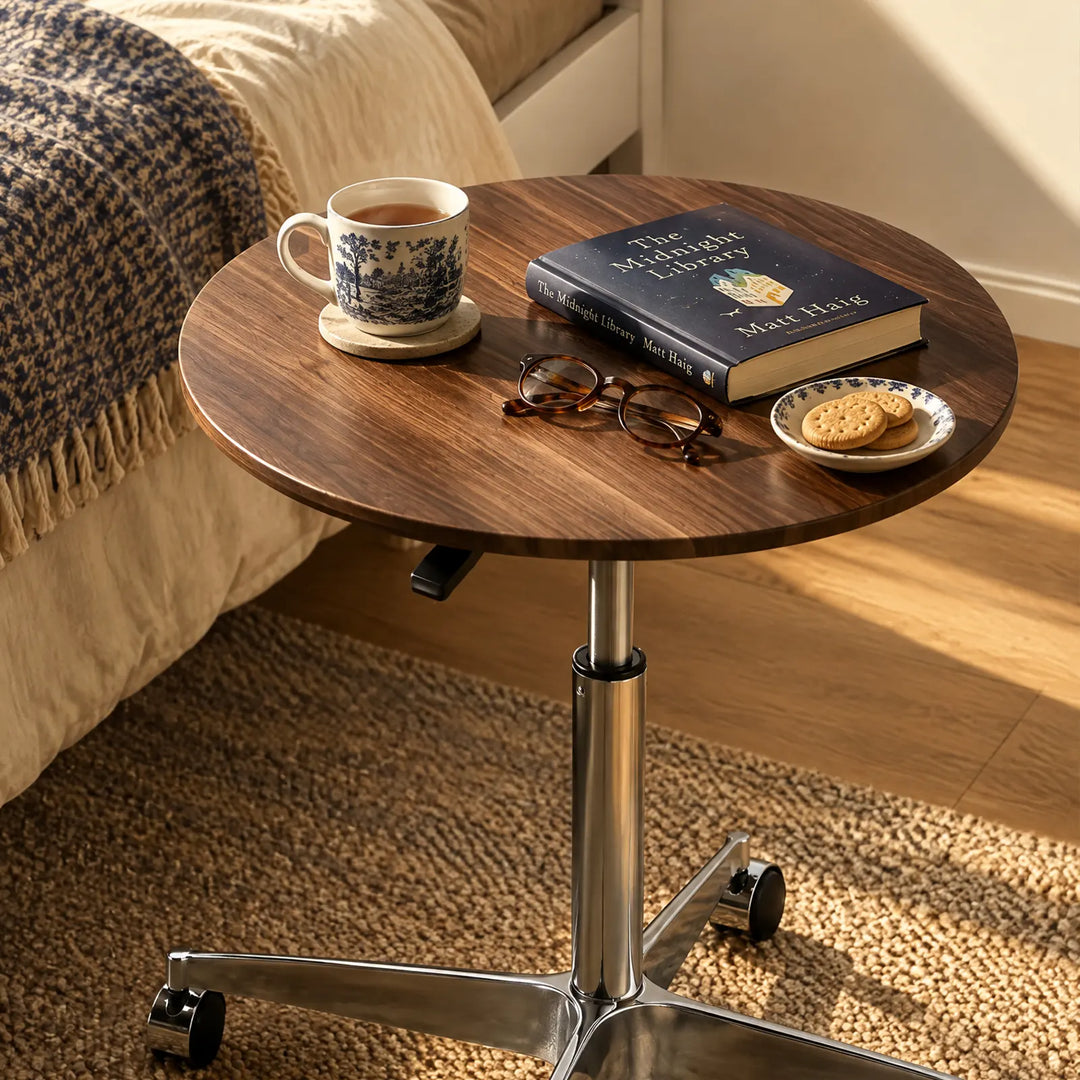 Showing a round side table with a walnut -ffect tabletop and wheels near a bed in a warm bedroom