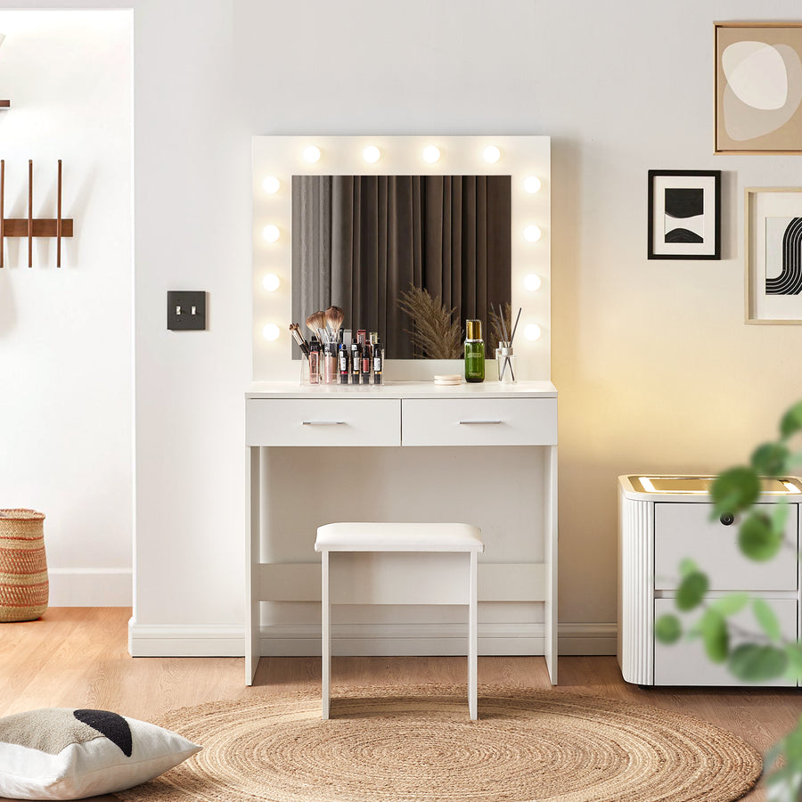 A Hollywood-style white vanity table with a mirror surrounded by bright bulb lights and a cushioned stool, placed in a modern bedroom with a bedside table beside it.